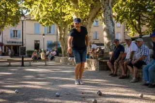 pétanque player mid-throw on a sunlit Provençal boulodrome, shaded by plane trees