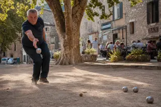 Player performing tir à la plombée on a shaded boulodrome with dust rising, warm Provençal light, and stone village buildings in the background.