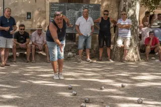 Pétanque player throwing boule on a shaded Provençal boulodrome, warm afternoon light, group of players watching, with shiny boules on a clay court and a hand-written scoreboard in the background.