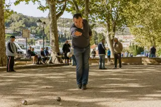 Pétanque player mid-throw on a shaded Provençal boulodrome with warm Mediterranean late afternoon light and casual atmosphere.