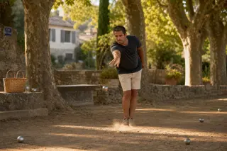 Pétanque player in a sunlit boule court under plane trees, mid-throw, with Provençal village ambiance and natural Mediterranean light.