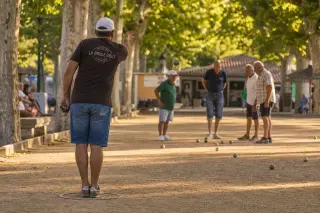 Petanque player throws boule as friends discuss positions near cochonnet on a shaded, sun-dappled boulodrome with plane trees, warm Mediterranean light.