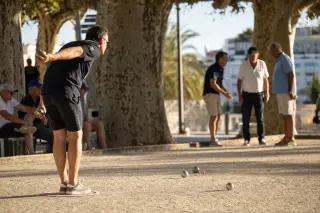 Pétanque player about to throw a boule on a shaded Cannes boulodrome under plane trees late in the afternoon, with others discussing near the cochonnet and a warm Mediterranean glow.