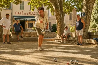 Pétanque player in action on a sunlit Provençal boulodrome with golden afternoon light, leafy shadows, and casual onlookers.