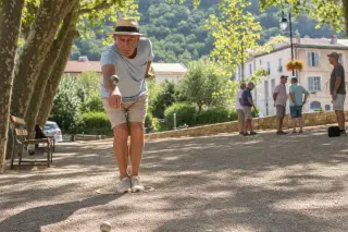 Pétanque player standing with feet together on a shaded gravel court, preparing to throw a boule under the warm Mediterranean sunlight filtered through plane trees.