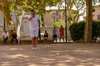 Pétanque player in sunlit Provençal boulodrome mid-throw under plane trees, late afternoon light, traditional atmosphere in Grasse