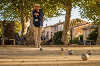 pétanque player points a boule on a sunlit, uneven Provençal boulodrome shaded by plane trees, evoking traditional Côte d’Azur atmosphere